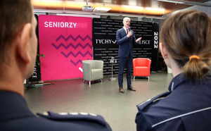 Mężczyzna przemawia przez mikrofon podczas konferencji, na pierwszym planie widoczne mundury policjantów.