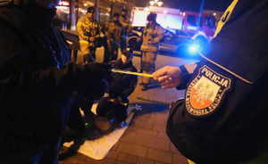 Na zdjęciu umundurowany policjant wręcza odblask pieszemu. Z tyłu strażacy i fantom do udzielania pierwszej pomocy.