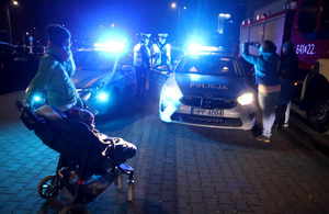 Policjanci stoją przy radiowozie z włączonymi sygnałami świetlnymi, kobieta robi zdjęcie, druga kobieta stoi z wózkiem z dzieckiem w środku.