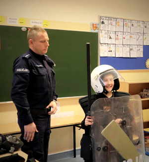 Umundurowany policjant stoi obok chłopiec ubrany w hełm, kamizelkę trzyma tarcze i pałkę.