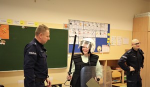 Dwóch umundurowanych policjantów w sali szkolnej, pomiędzy nimi chłopiec ubrany w hełm, kamizelkę, trzyma tarczę i pałkę.