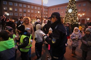 Na zdjęciu umundurowana policjantka rozdająca dzieciom Elementarze Bezpieczeństwa. Wokół policjantki dzieci i dorośli. W prawej częsci zdjęci choinka ze światełkami.