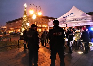 Na zdjęciu dwóch umundurowanych policjantów stojących tyłem. Przed nimi choinka ze światełkami oraz namiot z napisem Fundacja Rozwoju i Ekonomii Społecznej FRES. Pod namiotem umundurowana policjantka i inne osoby.