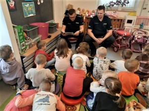 Na zdjęciu dwóch umundurowanych policjantów siedzących przed grupą dzieci. Dzieci siedzą przed policjantami na kolorowym dywanie. Wokół nich zabawki. Jeden z policjantów ma założone okulary i trzyma przed sobą książkę.