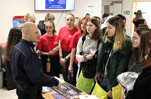 Na zdjęciu umundurowany policjant oraz grupa młodych ludzi, którzy z nim rozmawiają. Pomiędzy nimi stolik, na którym są rozłożone ulotki.