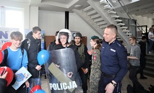 Na zdjęciu umundurowany policjant oraz grupa młodych ludzi. Jeden z chłopców ma założony sprzęt przeznaczony do zabezpieczania imprez masowych i trzyma w ręku policyjna tarczę.