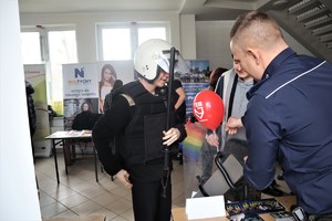 Na zdjęciu umundurowany policjant oraz dwóch uczniów. Jeden z nich ma założony sprzęt wykorzystywany przy zabezpieczeniu imprez masowych.