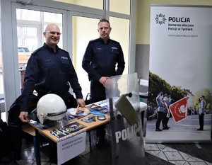 Na zdjęciu umundurowani policjanci przy stanowisku promocyjnym Policji. Przed stolikiem z ulotkami i plakatami stoi oparta tarcza. Na stoliku biały kask wykorzystywany przy zabezpieczaniu imprez masowych.