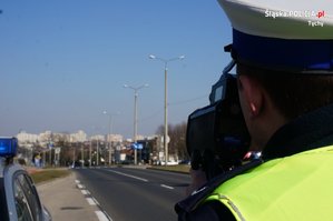 Na zdjęciu umundurowany policjant ruchu drogowego mierzący prędkość urządzeniem w rejonie przejścia dla pieszych. W oddali widać przejście dla pieszych i samochody osobowe.