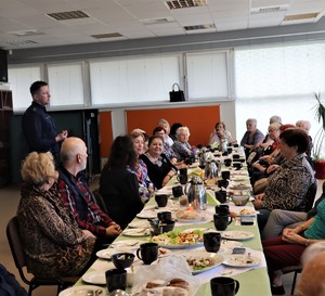 Na zdjęciu grupa seniorów siedzących przy stole. Obok nich stojący, umundurowany policjant.