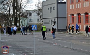 Na zdjęciu parking przed szkołą, na którym znajduje się tor przeszkód i miasteczko ruchu drogowego dla uczestników turnieju. Na śrdku zdjęcia umundurowany policjant w kamizelce odblaskowej, w oddali widać uczniów.