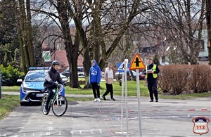 Na zdjęciu chłopiec jadący na rowerze. Na zdjęciu widoczny również umundurowany policjant w kamizelce odblaskowej oraz uczniowie.