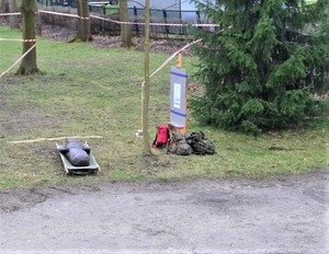 Na zdjęciu nosze z manekinem leżące na ziemi. Obok plecaki oraz odgrodzony taśmami tor.