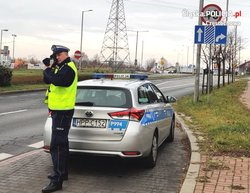 Na zdjęciu umundurowany policjant w kamizelce odblaskowej stojący obok radiowozu. Policjant trzyma w rękach urządzenie do mierzenia prędkości. W tle jezdnia i stojące obok niej znaki drogowe.