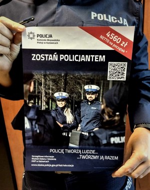 Na zdjęciu umundurowany policjant trzymający w rękach plakat zachęcający do pracy w Policji. Na plakacie umundurowani policjanci oraz napisy o treści: Zostań policjantem, Policję tworzą ludzie twórzmy ją razem