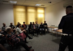 Na zdjęciu grupa osób siedzących na krzesłach. Przed nimi stoi umundurowany policjant.