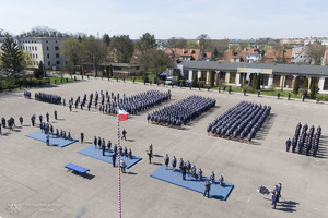 Na zdjęciu widoczny plac, na którym w równych kolumnach i szeregach ustawieni są umundurowali policjanci uczestniczący w uroczystości mianowania na oficerów Policji. W tle zdjęcia widoczne zabudowania. Przed policjantami widoczna wywieszona flaga Rzeczypospolitej Polskiej.