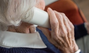 Na zdjęciu osoba starsza z siwymi włosami siedząca tyłem i trzymająca przy uchu słuchawkę telefonu stacjonarnego.