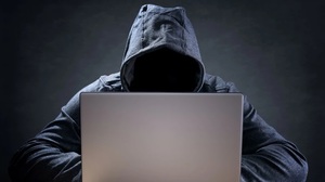 Na zdjęciu osoba w kapturze, z niewidoczną twarzą siedząca przed komputerem