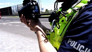 Na zdjęciu umundurowany policjant stojący przy drodze. Policjant trzyma w rękach urządzenie do pomiaru prędkości.