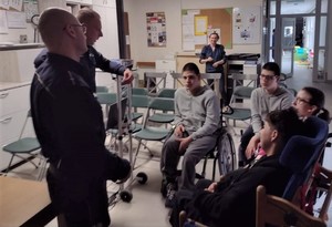 Na zdjęciu umundurowani policjanci stojący przed grupą młodych ludzi. Jeden z chłopców siedzi na wózku inwalidzkim, pozostali na krzesłach. W tle widać kobietę.