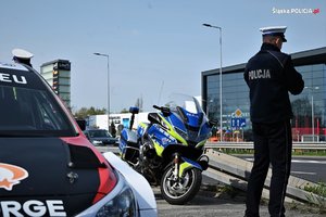 Na zdjęciu umundurowany policjant stojący przy drodze. Obok niego motocykl policyjny oraz widoczny fragment samochodu rajdowego. W tle zabudowania.