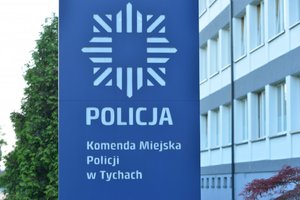 Na zdjęciu tablica informacyjna stojąca przed budynkiem komendy z napisem: Policja Komenda Miejska Policji w Tychach. Nad napisem logo Policji.
