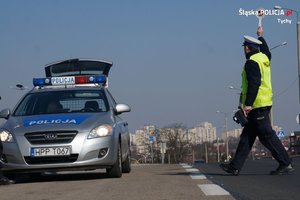 Na zdjęciu umundurowany policjant ruchu drogowego zatrzymujący pojazd do kontroli. Policjant stoi na jezdni z wyciągniętą ręką, w której trzyma tarczę do zatrzymywania pojazdów. Obok, na poboczu zaparkowany policyjny radiowóz.