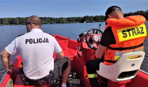 Na zdjęciu policjant i strażak, którzy siedzą w łódce płynącej po jeziorze. Funkcjonariusze odwróceni plecami.