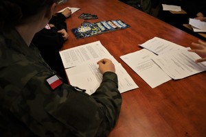 Na zdjęciu uczeń w szkolnym mundurze siedzący przy stole nad plikiem dokumentów. Na stole widoczne również gazety policyjne.