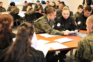 Na zdjęciu sala, na której przy stolikach siedzą uczniowie w szkolnych mundurach. Uczniowie przeglądają dokumenty.