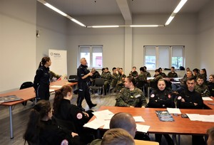 Na zdjęciu sala, na której przy stolikach siedzą uczniowie w szkolnych mundurach. Przed nimi dwoje umundurowanych policjantów.