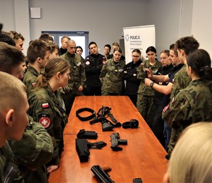 Na zdjęciu sala, na której przy stoliku zgromadzeni są uczniowie w szkolnych mundurach. Na stole rozłożone różne rodzaje broni.