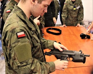 Na zdjęciu uczeń w szkolnym mundurze moro, który trzyma w dłoni broń policyjną. W tle inni uczniowie.