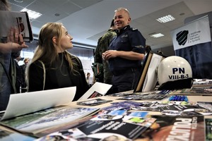 Na zdjęciu umundurowany policjant  oraz kobieta. Osoby te znajdują się obok stolika, na którym znajdują się policyjne gazety, ulotki i gadżety.