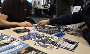 Na zdjęciu stolik z ulotkami oraz gadżetami promującymi pracę w Policji. Przy stoliku widać częściowo dwie siedzące osoby. Jedna z nich wskazuje palcem na ulotkę.