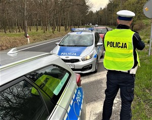 Na zdjęciu dwa policyjne radiowozy oraz umundurowany policjant ruchu drogowego, który kontroluje prędkość kierowców. W tle jezdnia i jadące nią samochody.