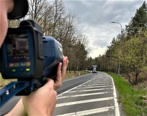 Na zdjęciu umundurowany policjant ruchu drogowego, który kontroluje prędkość kierowców. Policjant trzyma w rękach urządzenie do pomiaru prędkości. W tle jezdnia i jadące nią samochody.