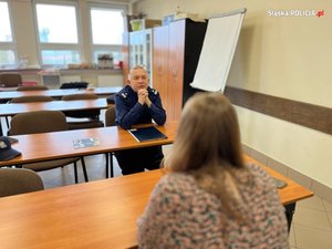 Na zdjęciu umundurowany policjant siedzący na krześle za stolikiem. Przed nim siedząca tyłem osoba.