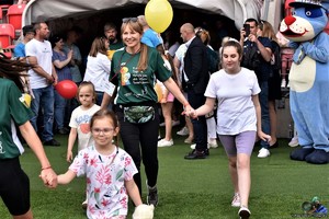 Na zdjęciu grupa osób, w tym dzieci, którzy wychodzą na murawę boiska. Za nimi po prawej stronie maskotka policyjnego psa Sznupka.