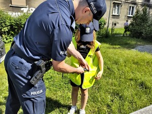 Na zdjęciu umundurowany policjant i dziewczynka. Policjant pomaga założyć dziewczynce kamizelkę odblaskową.