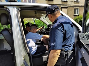 Na zdjęciu umundurowany policjant stojący obok oznakowanego radiowozu policyjnego. Z kierownicą i na przednim siedzeniu pasażera siedzi dwóch chłopców.