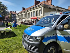 Na zdjęciu oznakowany policyjny radiowóz, obok którego znajdują się dzieci i policjanci. W tle zielony skwer i osoby uczestniczące w pikniku rodzinnym. Widać parasole i balony.
