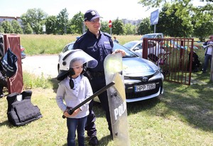 Na zdjęciu umundurowany policjant oraz chłopiec z założoną kamizelką i kaskiem. Chłopiec trzyma  tarczę i policyjną pałkę. Za nimi oznakowany radiowóz. .