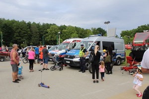 Na zdjęciu stojące w rzędzie pojazdy uprzywilejowane. Wśród nich motocykl policyjny, radiowóz i karetka Pogotowia Ratunkowego. Przed motocyklem policjant umundurowany Ruchu Drogowego oraz osoby uczestniczące w pikniku.