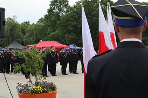 Na zdjęciu stojący tyłem strażak. Przed nim flaga Rzeczypospolitej Polskiej. W tle strażacy stojący na placu apelowym.