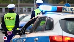Na zdjęciu dwóch umundurowanych policjantów Wydziału Ruchu Drogowego stojących obok radiowozu. W tle droga z samochodami.