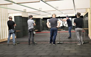Na zdjęciu grupa czterech mężczyzn stojących tyłem. Przed nimi mężczyzna z uniesioną bronią. W tle widać tarcze do strzelań.