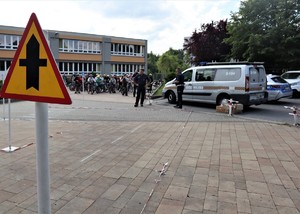 Na zdjęciu grupa dzieci. Niektóre z nich mają stoją na rowerach i mają założone kaski rowerowe. Przed nimi umundurowany policjant oraz dwóch funkcjonariuszy Straży Miejskiej.