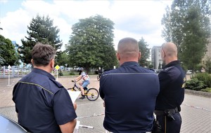 Na zdjęciu umundurowany policjant oraz funkcjonariusze Straży Miejskiej. W tle dzieci jeżdżące na rowerach.